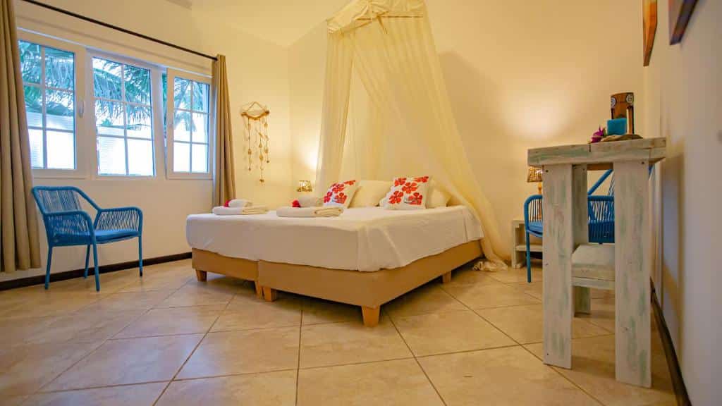 Quarto do Hibiscus Beach House com uma cama de casal, travesseiros florais, duas cadeiras azuis, uma pequena escrivaninha e janelas grandes com cortinas.