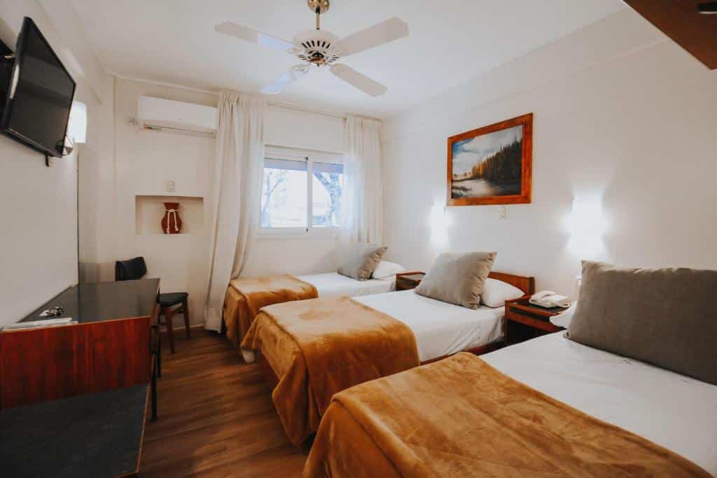 Três camas de solteiro do Hotel Nutibara com cobertores marrons em um quarto de hotel com escrivaninha, TV, ventilador de teto e janela com cortinas brancas.