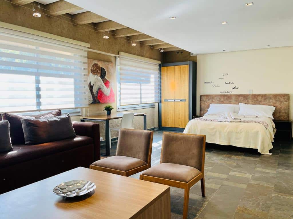 Quarto espaçoso da La Sofia Apart & Wines com uma cama de casal, sofá marrom, duas cadeiras, janelas grandes e arte na parede acima da cabeceira.