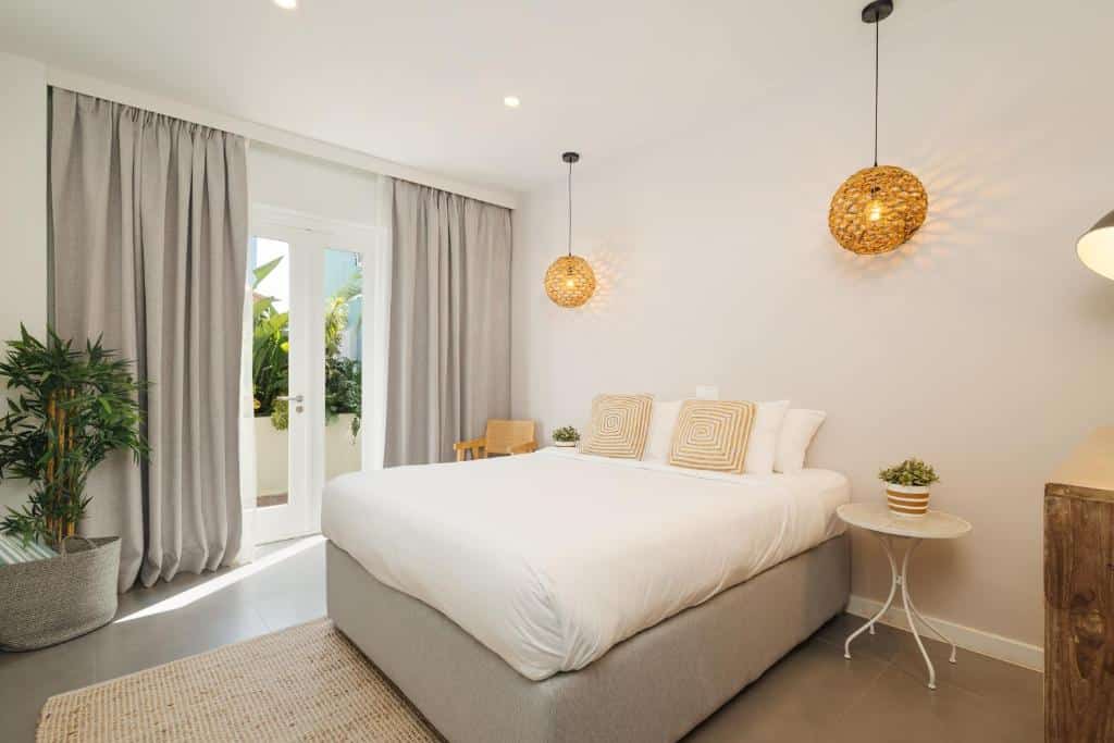 Um quarto do Pietermaai Oasis com uma cama de casal, duas luminárias penduradas, grandes cortinas cinza, plantas e uma porta que dá para um jardim.