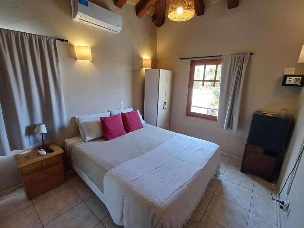 Uma cama bem arrumada da Posada Cavieres Wine Farm com roupa de cama branca e travesseiros vermelhos em um quarto pequeno com guarda-roupa, janela e vigas de madeira no teto.