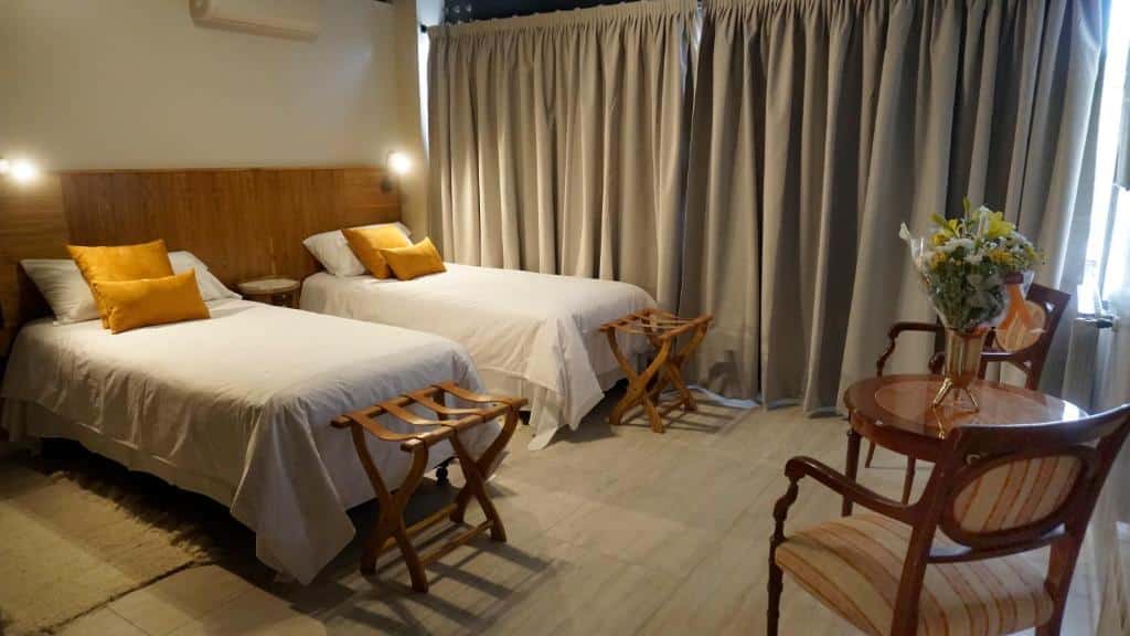 Quarto do 718 Rufino Petit Hotel com duas camas, cabeceiras de madeira, travesseiros amarelos, uma mesa redonda com flores e duas cadeiras de madeira.