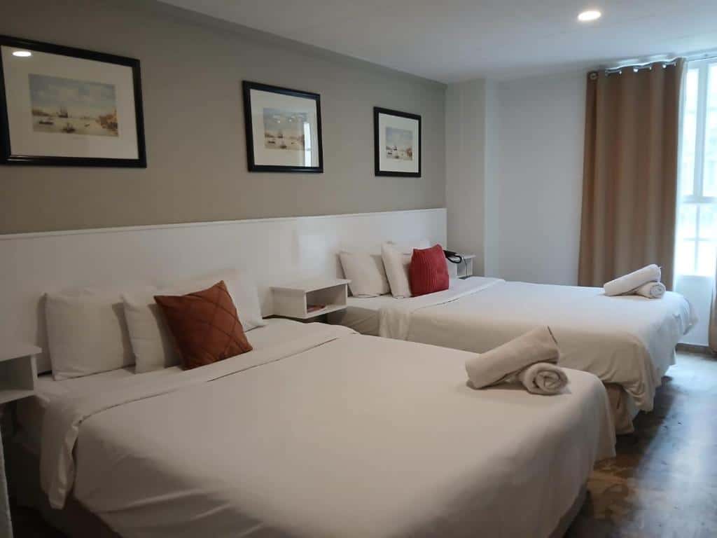 Quarto do San Marco Hotel Curacao & Casino com duas camas de casal, roupa de cama branca, travesseiros decorativos, arte emoldurada e uma janela com cortinas bege.