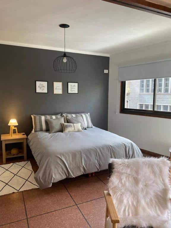 Um quarto do Terrazas de Rondeau com uma parede cinza de destaque, cama com roupa de cama cinza, criado-mudo, luminária, janela e decoração de parede acima da cama.