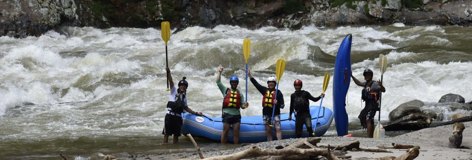 Cinco pessoas com coletes salva-vidas e capacetes estão ao lado de um bote inflável azul segurando remos perto de um rio em movimento rápido. Imagem para ilustrar post sobre o rafting de nível avançado no rio Samaná.