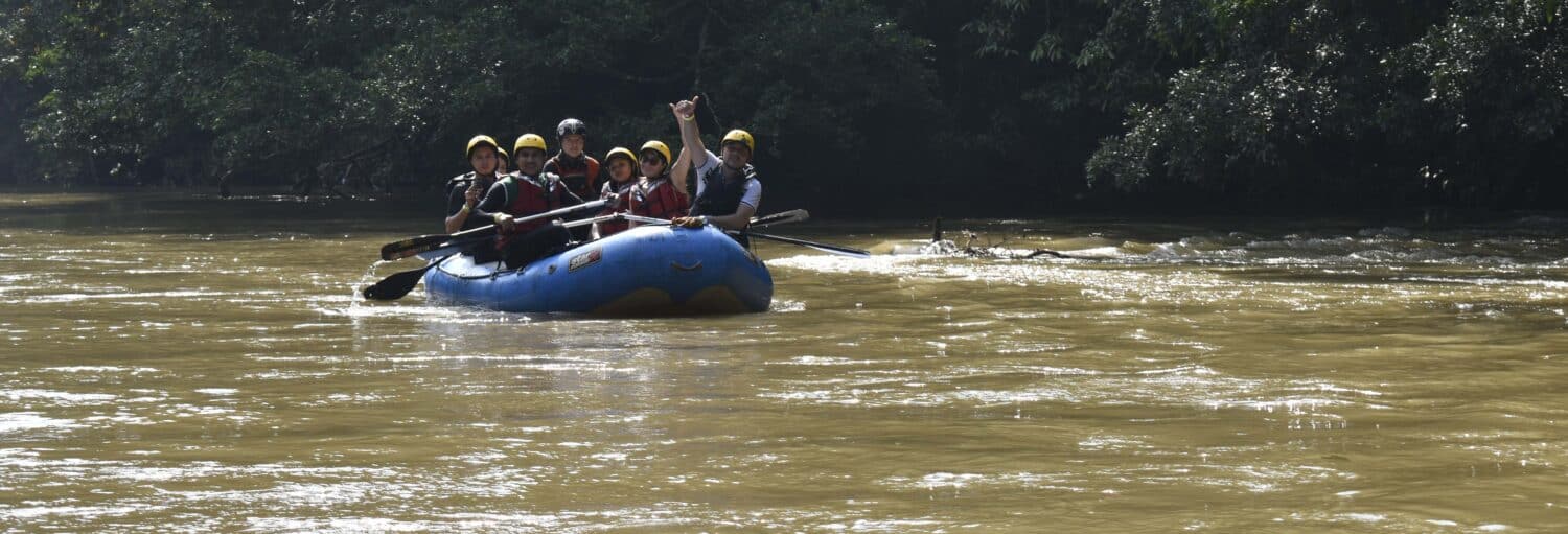 Um grupo de pessoas usando capacetes e coletes salva-vidas rema um bote inflável azul em um rio lamacento perto de árvores. Imagem para ilustrar post sobre Rafing no rio Claro.