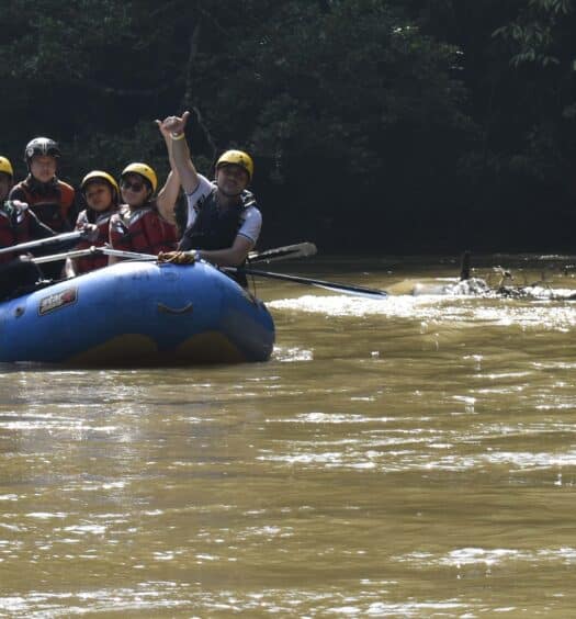 Um grupo de pessoas usando capacetes e coletes salva-vidas rema um bote inflável azul em um rio lamacento perto de árvores. Imagem para ilustrar post sobre Rafing no rio Claro.