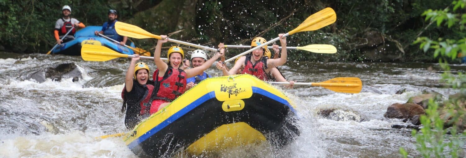 Um grupo de pessoas usando capacetes e coletes salva-vidas praticando rafting em um rio, com remos levantados e respingos de água. - Rafting no rio Paranhana
