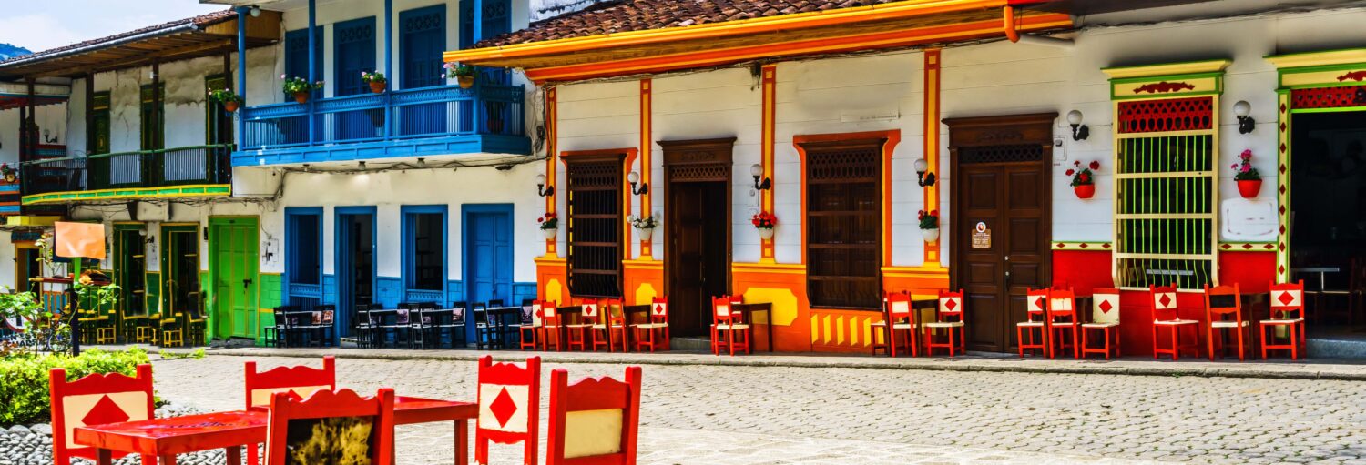 Cadeiras e mesas coloridas se alinham do lado de fora de edifícios vibrantes e multicoloridos ao longo de uma rua de paralelepípedos. Imagem para ilustrar post sobre a excursão a Jardín.