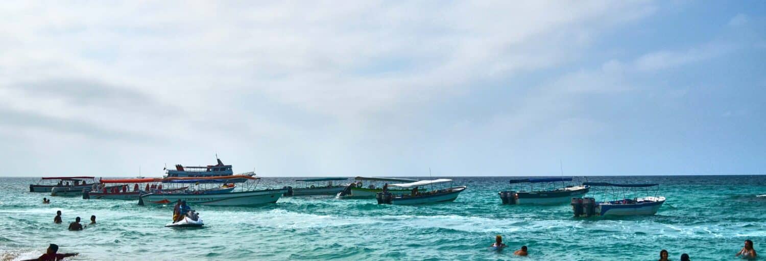As pessoas nadam em águas azul-turquesa perto da costa, com vários barcos flutuando no mar sob um céu parcialmente nublado. Imagem para ilustrar post sobre snorkel na península de Barú.