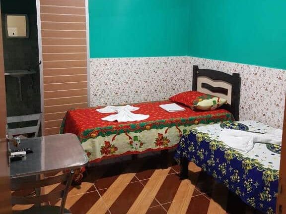 Um quarto pequeno da pousada pouso feliz com duas camas de solteiro, uma escrivaninha e uma cadeira de metal, paredes azul-petróleo e uma porta aberta para o banheiro.