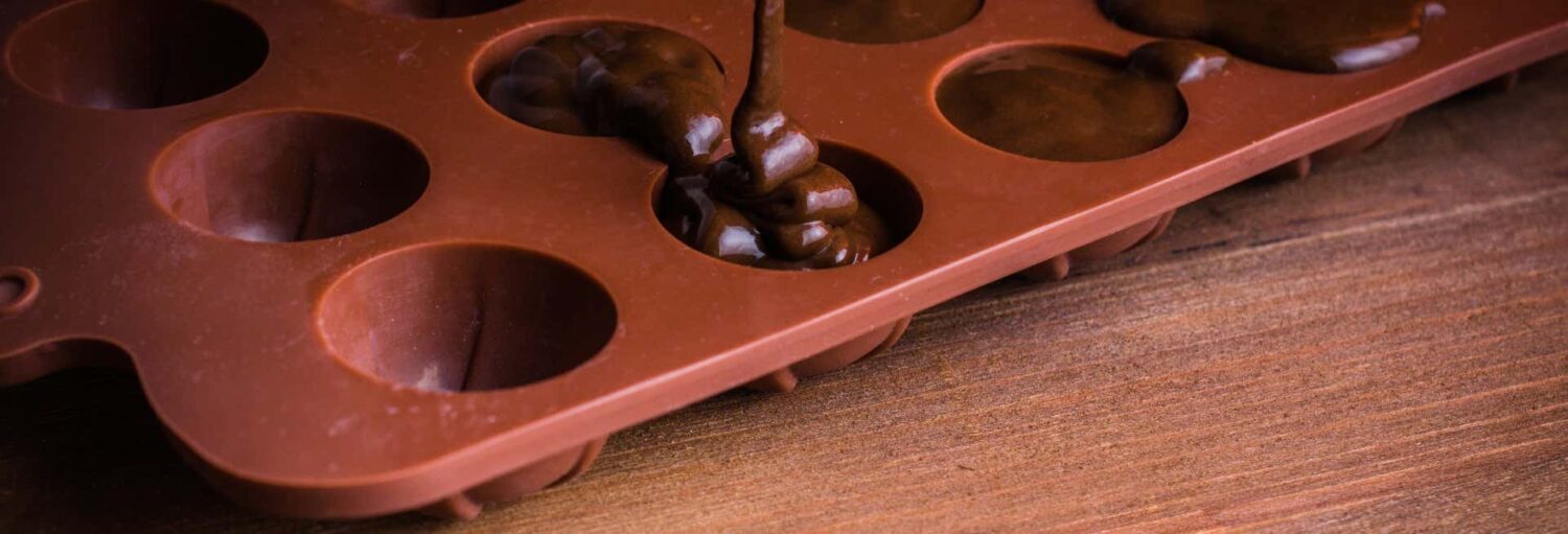 O chocolate está sendo despejado em um molde de silicone marrom com várias cavidades vazias e parcialmente preenchidas em uma superfície de madeira. Imagem para ilustrar post sobre a oficina de chocolate em Cartagena das Índias.