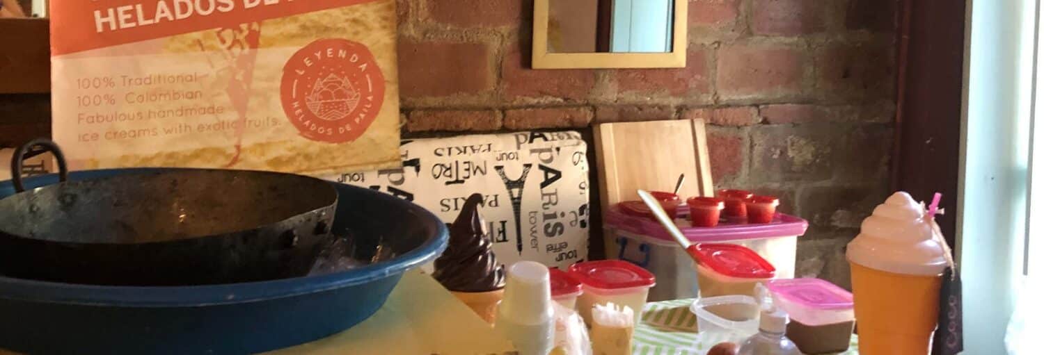 Mesa com recipientes, uma casquinha de sorvete e uma placa anunciando sorvete artesanal colombiano contra uma parede de tijolos. Imagem para ilustrar post sobre a oficina e degustação de sorvete de paila.