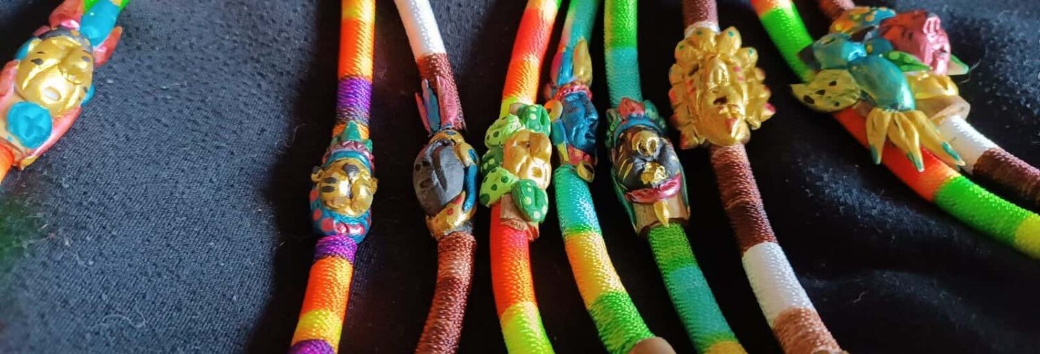Pulseiras de tecido colorido com máscaras pequenas e detalhadas e figuras de animais exibidas em um fundo de tecido preto. A imagem ilustra o post sobre Oficina de medicina ancestral indígena em Bogotá.