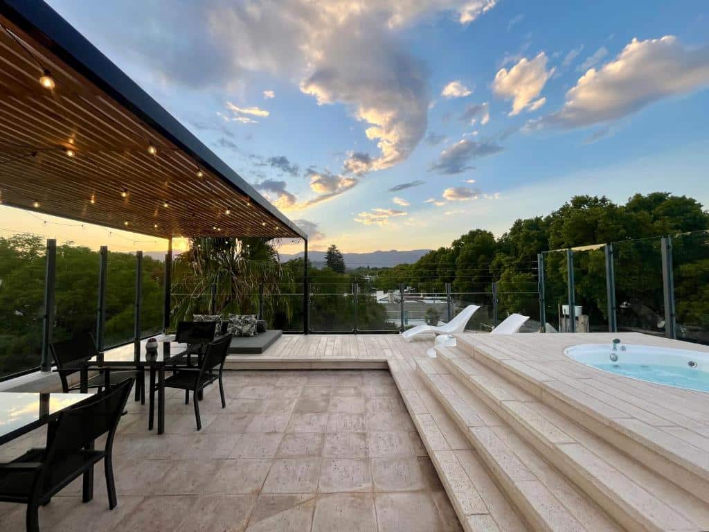 Pátio na cobertura da La Sofia Apart & Wines com mesas, cadeiras, espreguiçadeiras, banheira de hidromassagem, luzes de corda e vista para as árvores e montanhas ao pôr do sol.
