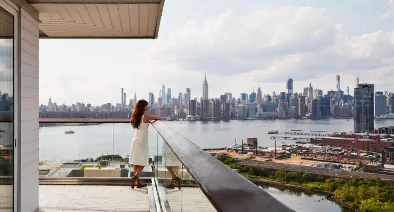 Uma mulher está em na varanda do hotel The William Vale, em Williamsburg, com vista para um rio e para o horizonte da cidade de Nova York em um dia parcialmente nublado. A foto é capa do post de onde ficar no Brooklyn em Nova York.