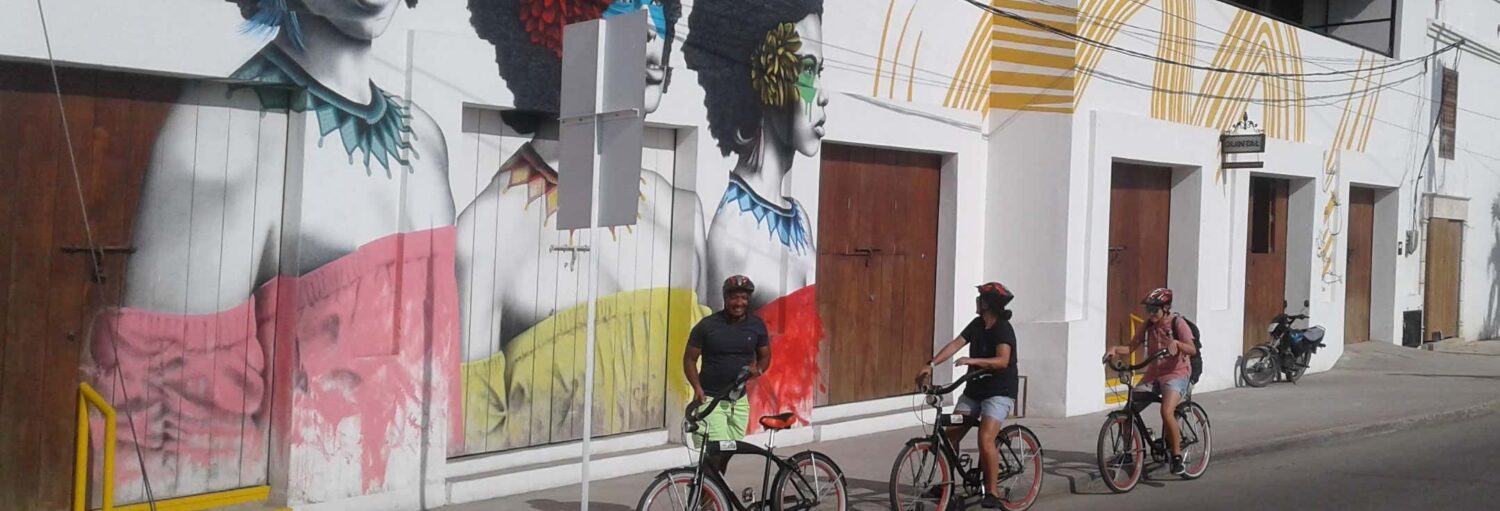 Três pessoas passam de bicicleta por um mural colorido de rostos de mulheres pintados em um prédio branco com portas de madeira. Imagem para ilustrar post sobre o tour de bicicleta por Cartagena das Índias.