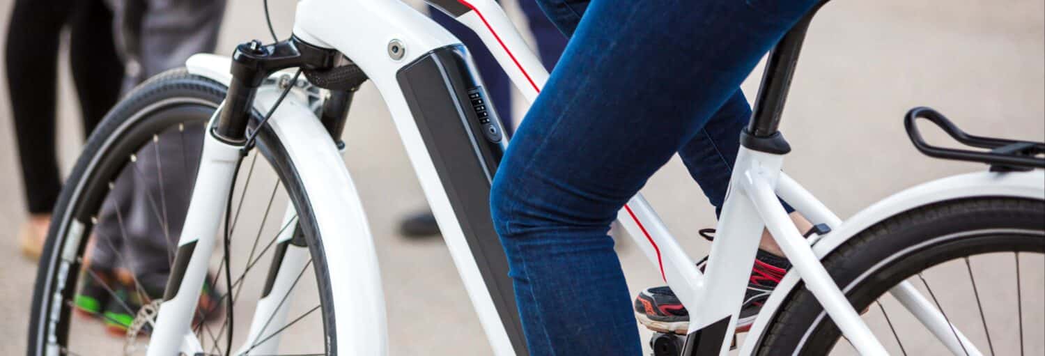 Pessoa de jeans e tênis andando em uma bicicleta elétrica branca em uma superfície pavimentada; outras pessoas visíveis ao fundo. Representa o post sobre o Tour de bicicleta elétrica por Atenas