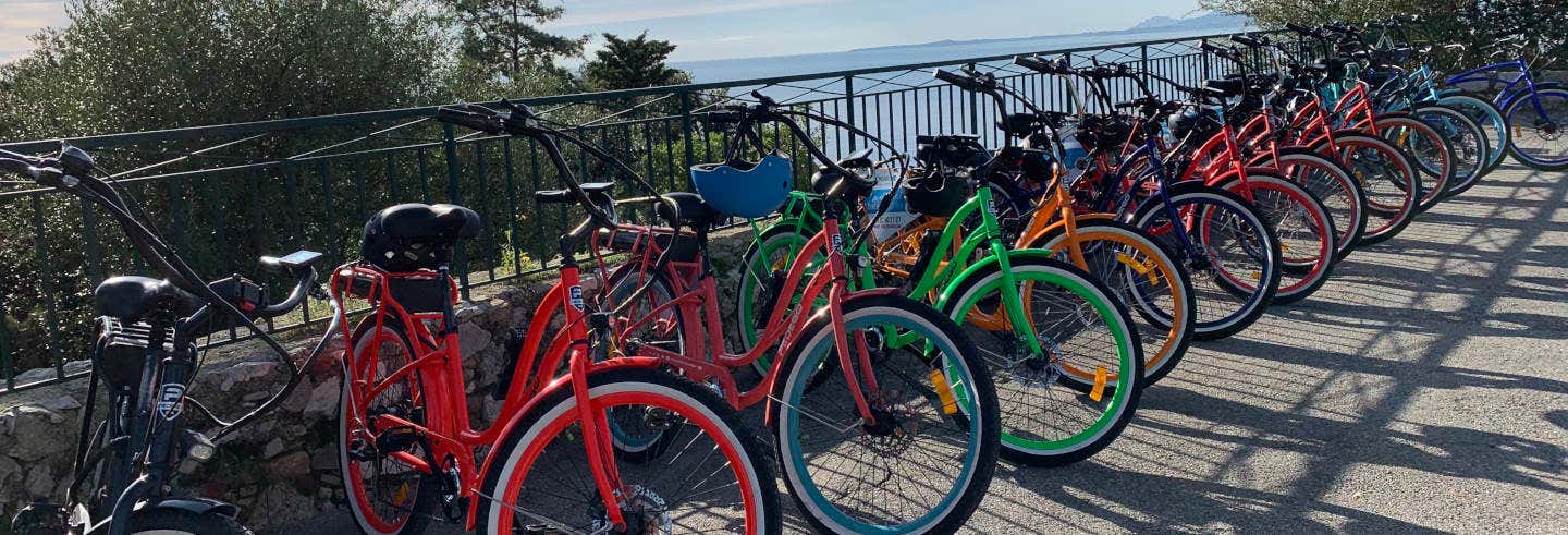 Uma fileira de bicicletas coloridas, a maioria vermelha com algumas verdes, amarelas e azuis, alinhadas em uma grade com vista para a água. - Tour de bicicleta elétrica por Nice