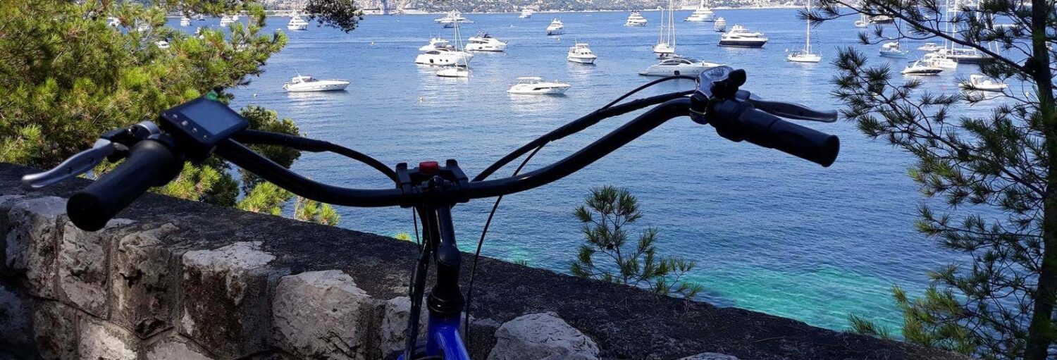 Guidão de bicicleta em primeiro plano, com vista para um muro de pedra, mar azul e barcos ancorados com árvores em ambos os lados. - Ilustra o Tour de bicicleta elétrica por Villefranche-sur-Mer