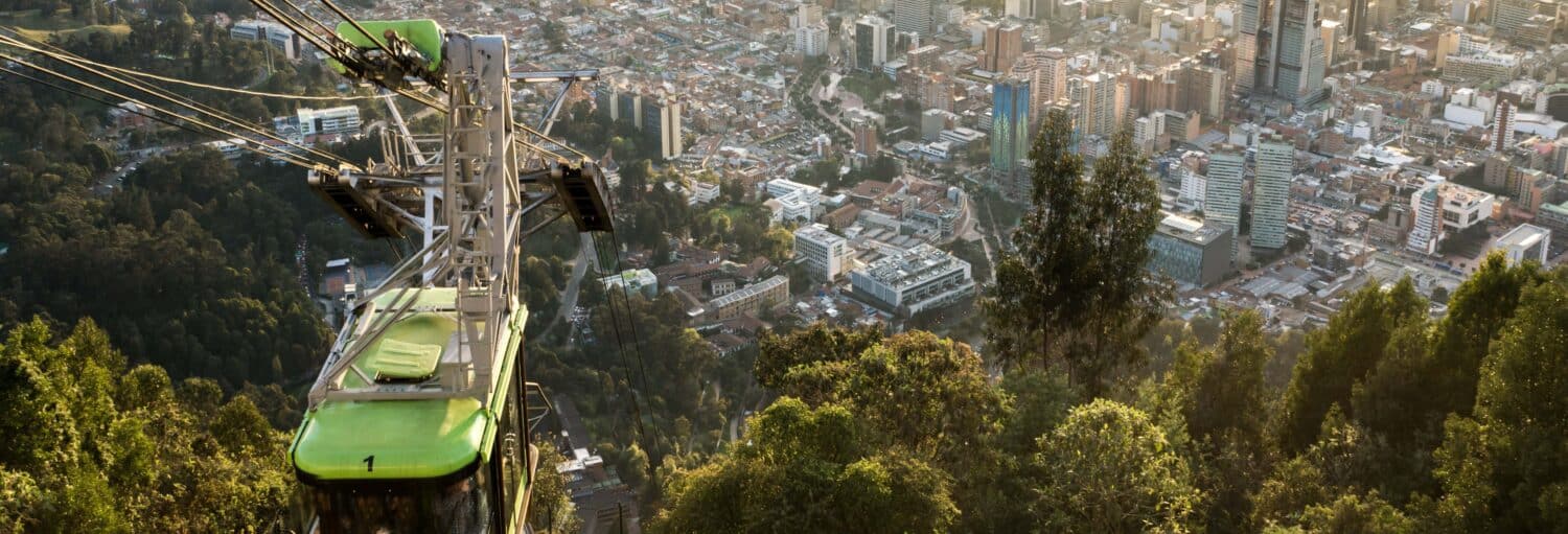 Um teleférico verde sobe sobre colinas arborizadas com uma paisagem urbana espalhada ao fundo. A imagem ilustra o post sobre Tour completo de Bogotá com ingressos.