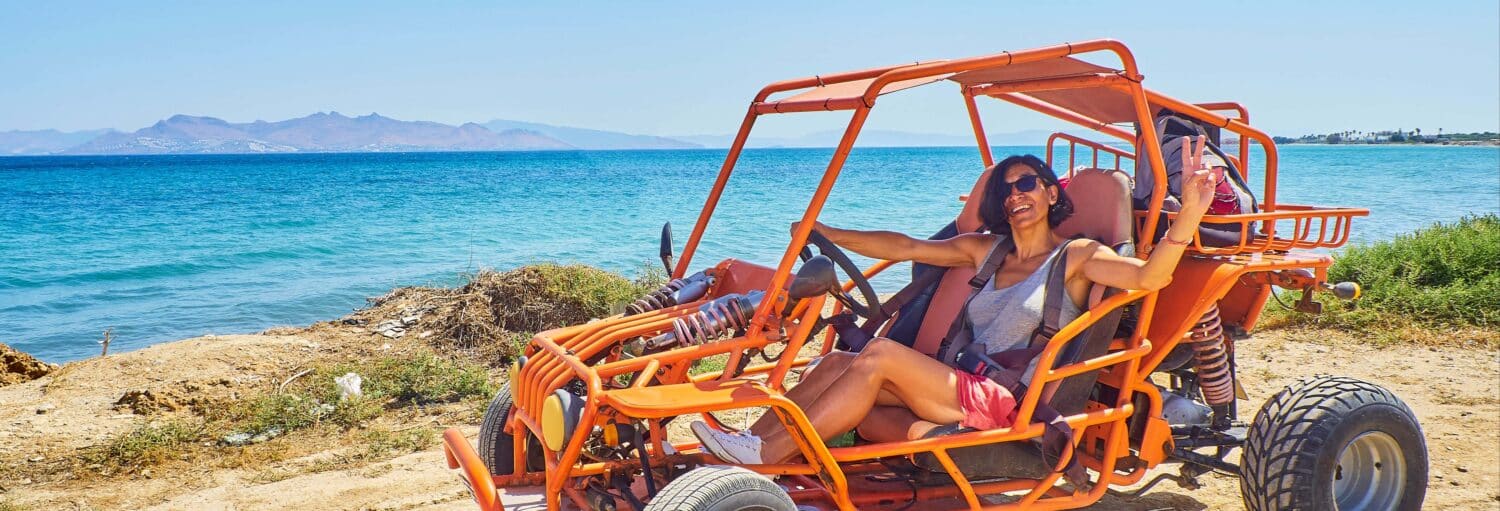 Duas pessoas sentadas em um buggy de dunas laranja estacionado perto de um mar azul, com montanhas visíveis no fundo distante. Imagem para ilustrar post sobre o tour de buggy pelas praias de Cartagena.