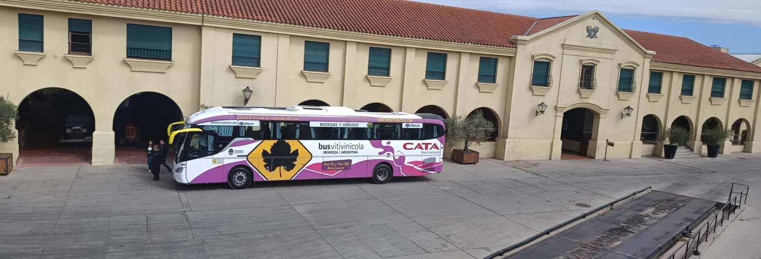 Um ônibus urbano branco e roxo está estacionado do lado de fora de um grande edifício bege com persianas verdes e um telhado de telhas vermelhas do Tour pelo pelo Caminho do Vinho das Grandes Lendas de Maipú