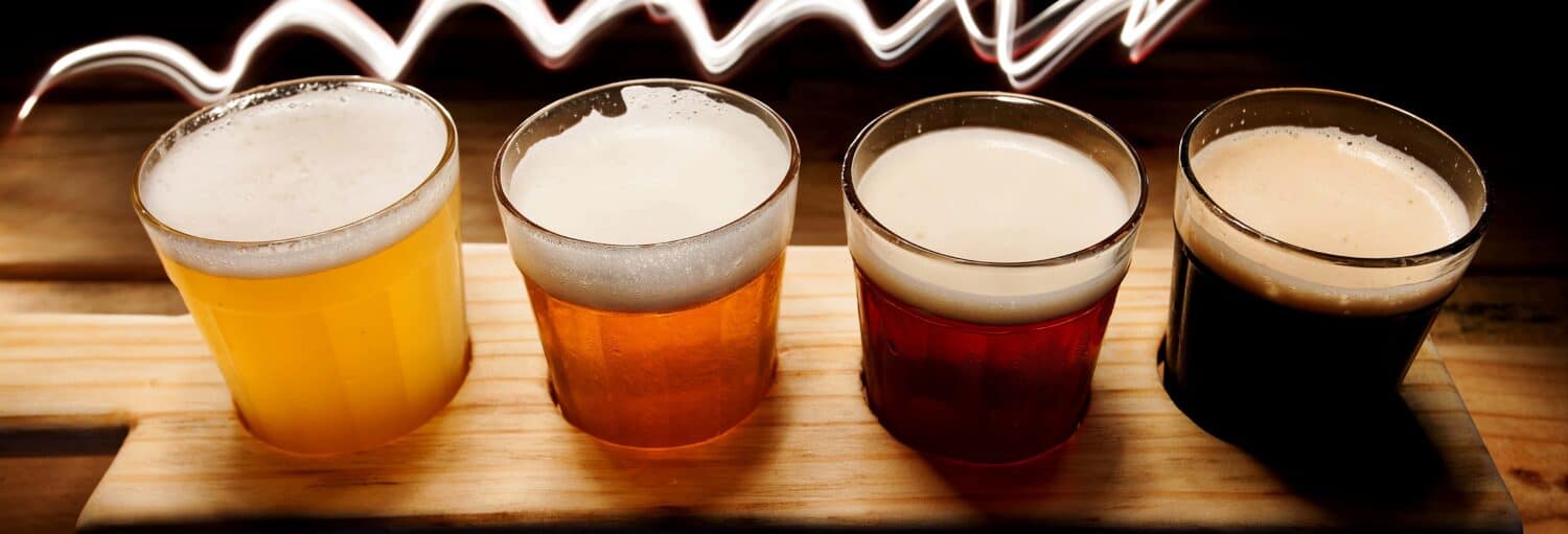 Quatro copos de cervejas de cores diferentes em um quadro de madeira, com um padrão ondulado de luz branca ao fundo. a imagem ilustra o post sobre Tour da cerveja artesanal por Bogotá.