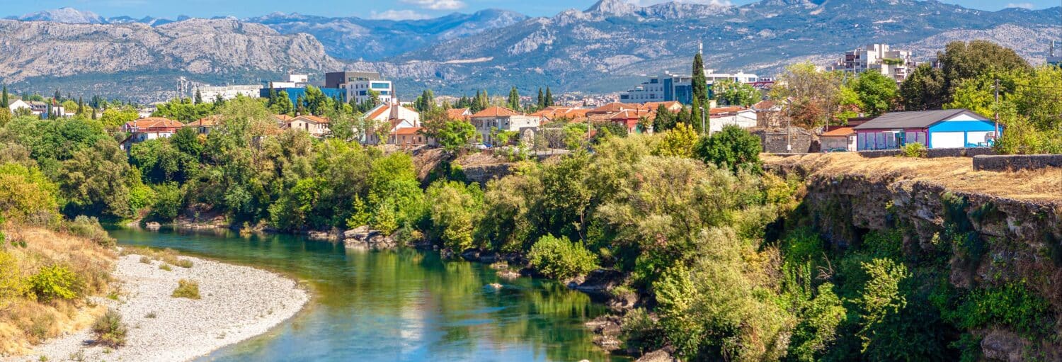Um rio passa por árvores e edifícios com montanhas ao fundo, sob um céu azul com nuvens dispersas, para representar Tour pela costa de Podgorica.