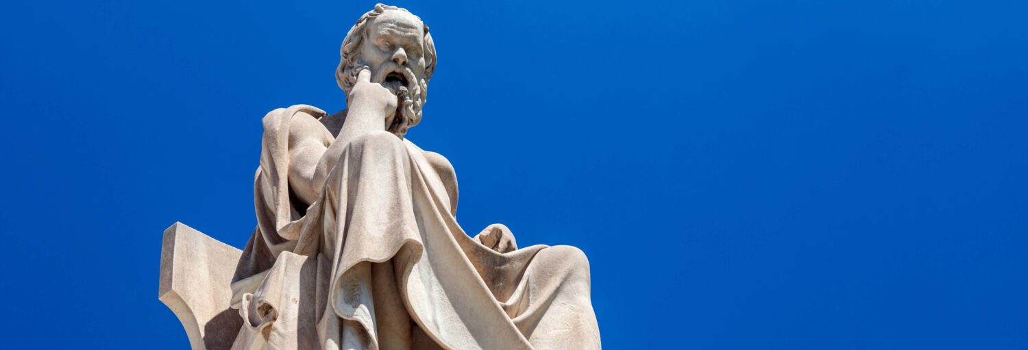Estátua de mármore de um homem barbudo sentado com vestes contra um céu azul claro, capturada de um ângulo baixo. A imagem ilustra o post sobre Tour da filosofia grega por Atenas.