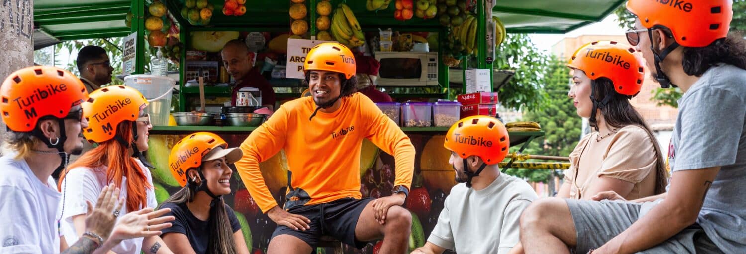Um grupo de pessoas usando capacetes laranja senta e conversa perto de uma barraca de frutas com um vendedor e várias frutas visíveis. Imagem para ilustrar post sobre o tour gastronômico de bicicleta elétrica por Medellín.