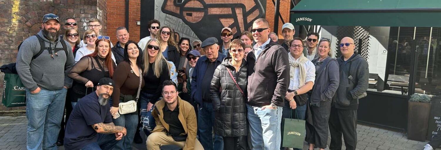 Um grupo de pessoas posando para uma foto ao ar livre perto de um mural e de um toldo verde da Jameson. A foto é capa do post do Tour pela Guinness Storehouse + Destilaria Jameson.
