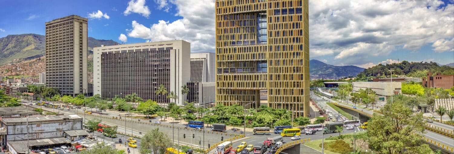 Vista panorâmica de uma cidade com altos edifícios de escritórios, ruas movimentadas, árvores e montanhas ao fundo, sob um céu parcialmente nublado. Imagem para ilustrar post sobre o tour completo por Medellín.