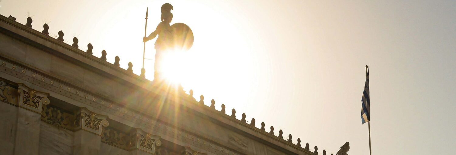 O sol brilha atrás de uma estátua de um guerreiro no telhado de um prédio, com um mastro e uma bandeira grega visíveis à direita. A imagem ilustra o post sobre Tour noturno por Atenas.