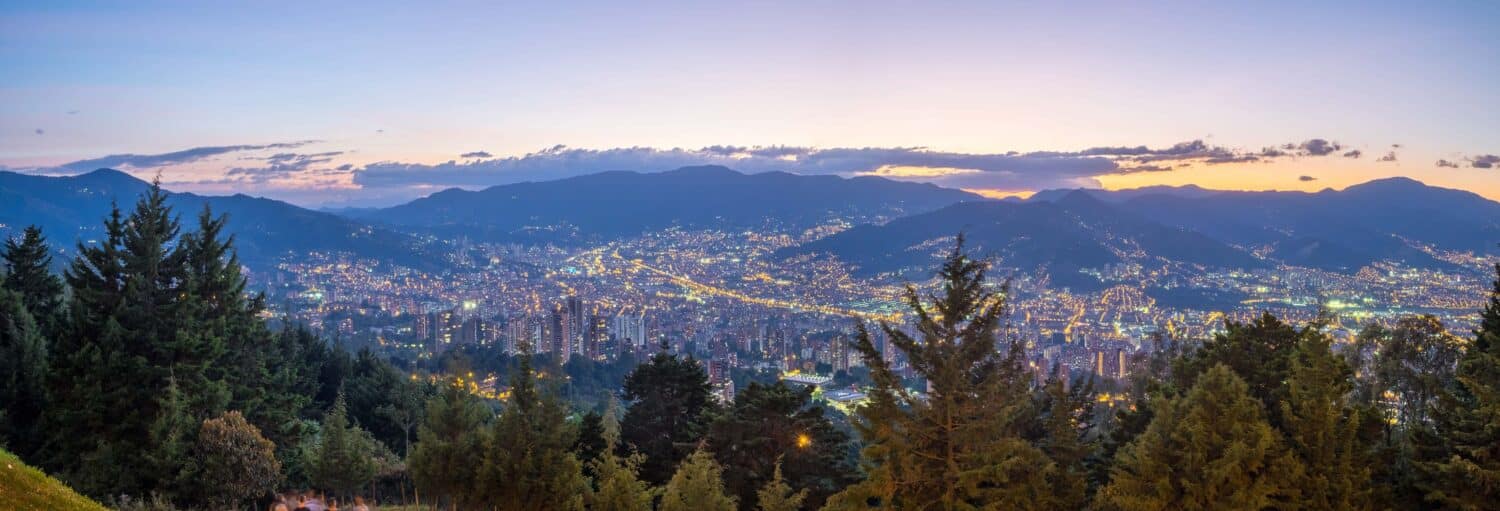 Vista panorâmica de uma cidade com prédios altos, árvores em primeiro plano e montanhas ao fundo ao anoitecer. Imagem para ilustrar post sobre o tour pelos mirantes de Medellín.