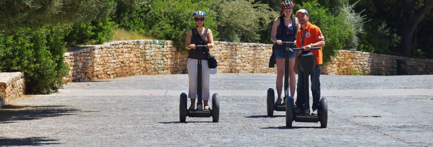Três pessoas usando capacetes andam de Segways em um caminho de paralelepípedos cercado por muros de pedra e vegetação. A imagem ilustra o post sobre Tour de segway por Atenas.
