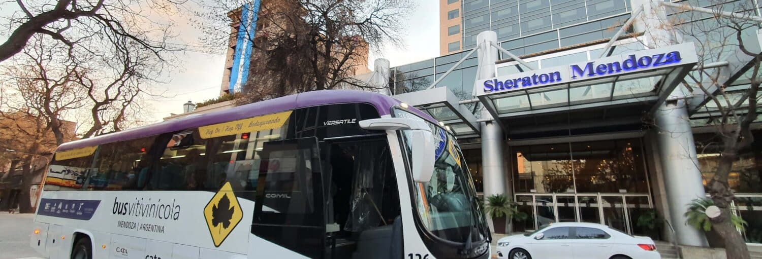 Um grande ônibus está estacionado em frente à entrada do Sheraton Mendoza Hotel, em uma rua da cidade com árvores e prédios altos durante o Tour pela zona vitivinícola de Maipú