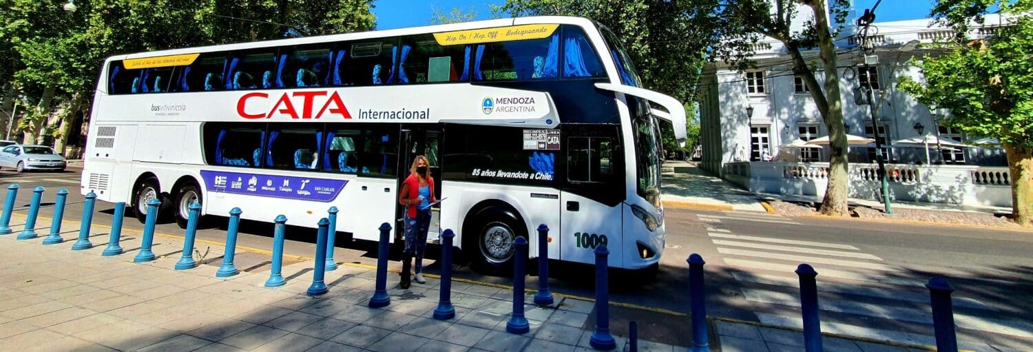 Um ônibus CATA Internacional de dois andares está estacionado em uma rua da cidade com árvores e uma mulher caminhando nas proximidades durante o Tour pelo sul da zona vitivinícola do Vale do Uco