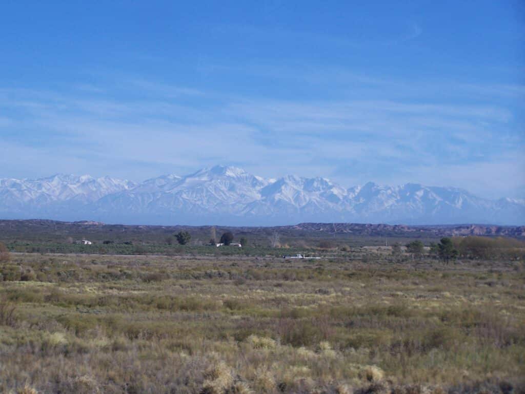 Montanhas cobertas de neve à distância, atrás de uma planície gramada e plana, sob um céu azul com nuvens claras.