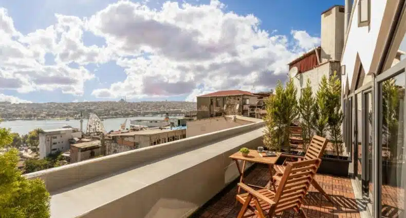 Um terraço ensolarado com cadeiras e mesas de madeira tem vista para uma paisagem urbana e um corpo d'água sob um céu parcialmente nublado. Imagem do Doruk Palas Hotel para ilustrar post sobre onde ficar em Istambul.