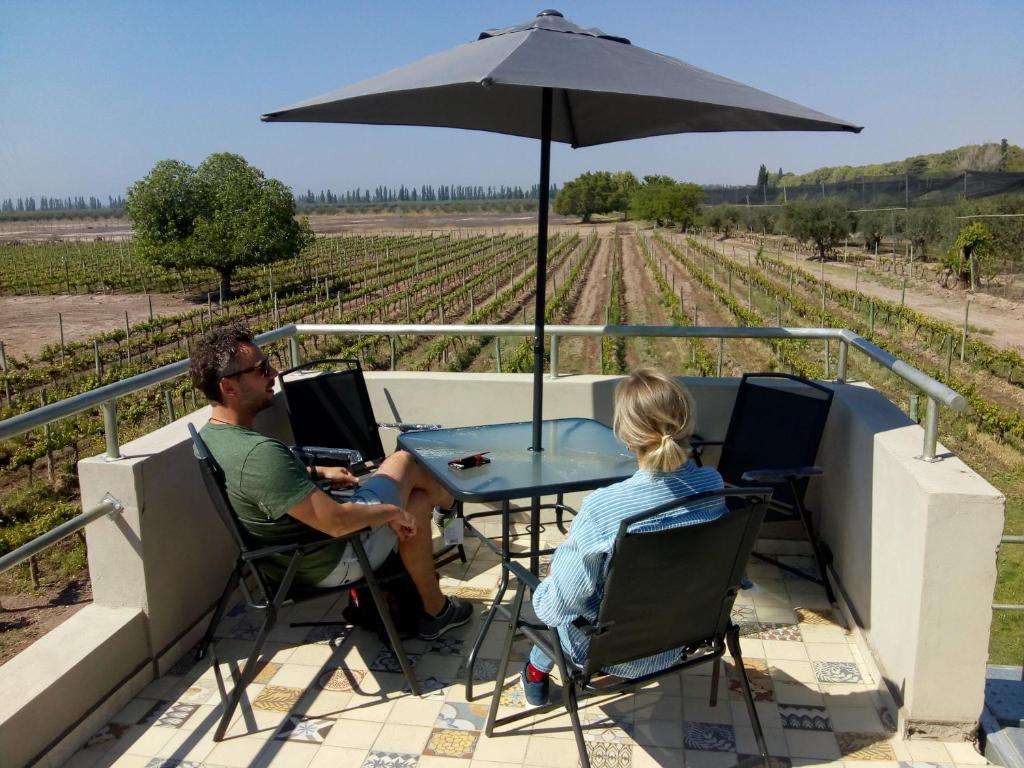 Duas pessoas sentam-se em uma mesa sob um guarda-sol em um pátio da Posada Cavieres Wine Farm com vista para um grande vinhedo em um dia ensolarado. Imagem para ilustrar post sobre onde ficar em Mendoza.