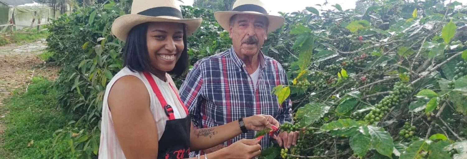 Duas pessoas de chapéu em meio a plantas de café em uma Visita à fazenda cafeeira El Edén, sorrindo com cerejas de café.