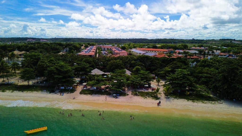 Vista aérea de uma praia de areia com água verde, pessoas nadando, árvores e edifícios ao fundo sob um céu nublado.