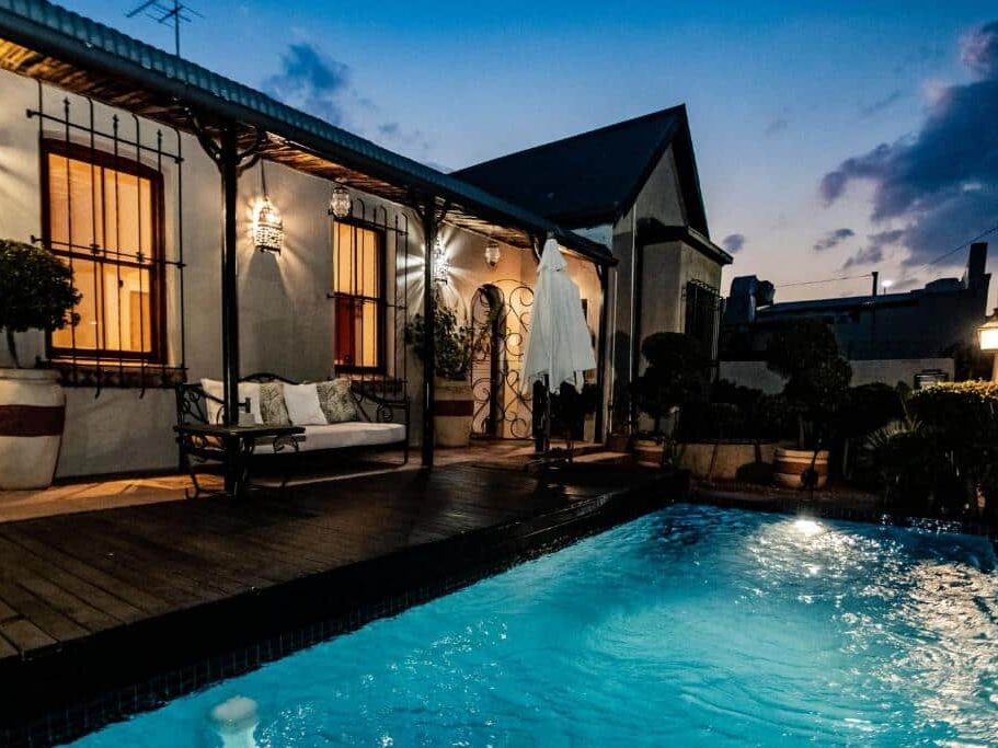 Uma piscina pequena e iluminada no 84 on Fourth Guest House, ao lado da casa com varanda, banco e vasos de plantas ao anoitecer. A foto é capa do post de onde ficar em Joanesburgo.