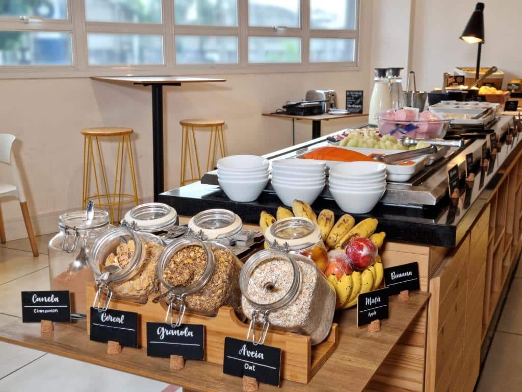 Um bufê de café da manhã do B&B HOTEL Rio de Janeiro Norte com potes de cereais, frutas, tigelas, pratos e vários itens alimentares em um balcão de madeira em uma sala iluminada.