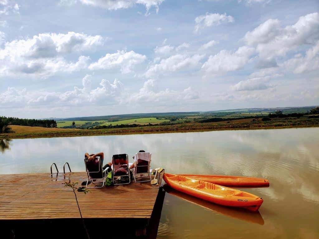 Duas pessoas relaxam em espreguiçadeiras em uma doca em um lago calmo com dois caiaques laranja, sob um céu parcialmente nublado.