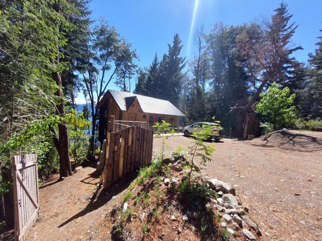 Uma pequena casa de madeira fica entre as árvores, com um carro estacionado nas proximidades, em uma clareira de terra, sob um céu azul claro.