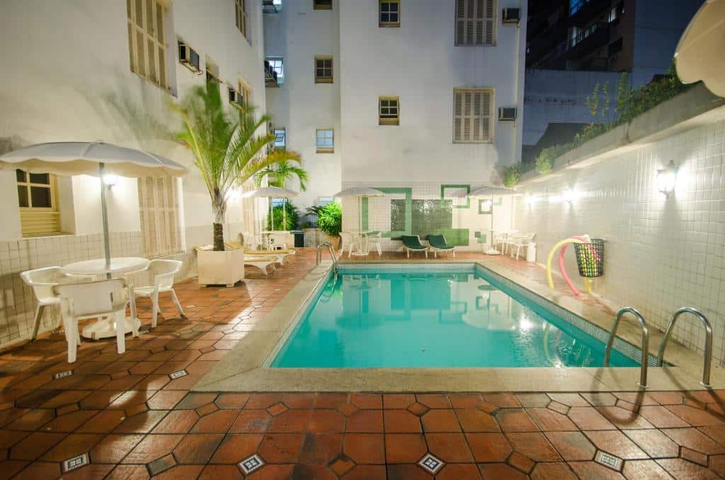 Piscina retangular ao ar livre cercada por mesas, cadeiras, plantas e paredes de azulejos brancos em um pátio urbano do Hotel Monte Alegre à noite.