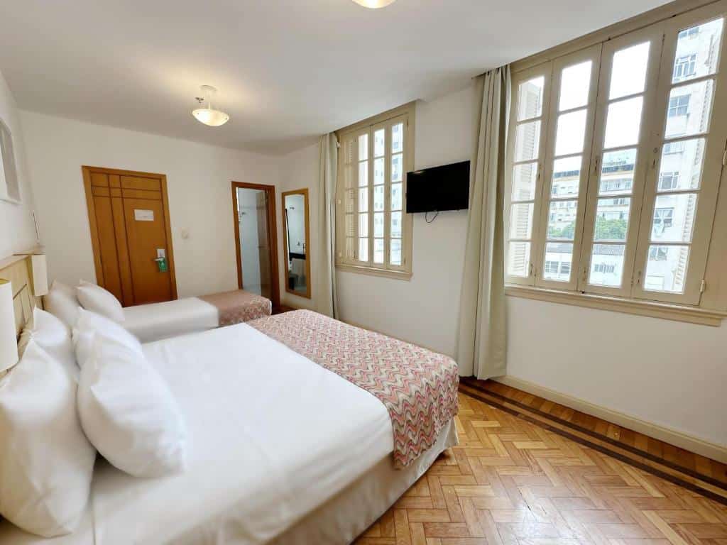 Quarto do Hotel Monte Alegre com uma cama de casal e uma cama de solteiro, piso de madeira, janelas grandes, TV na parede, porta e espelho. Essa é a uma das opções de onde ficar no Rio de Janeiro.