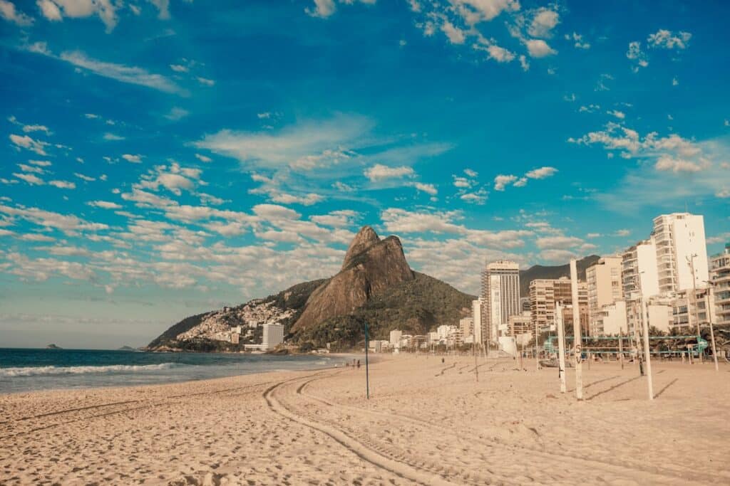 Uma praia de areia com trilhas, prédios urbanos e uma montanha sob um céu azul parcialmente nublado.
